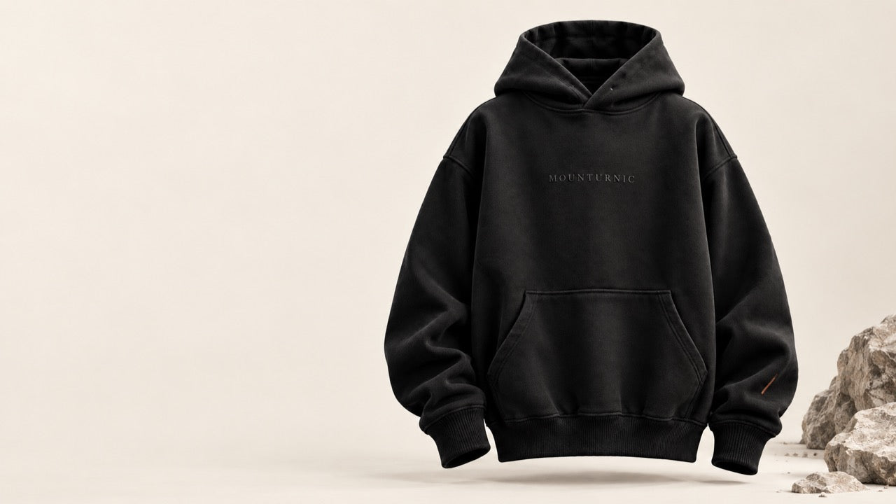 The black hoodie standing against a warm ivory background beside pale rocks.
